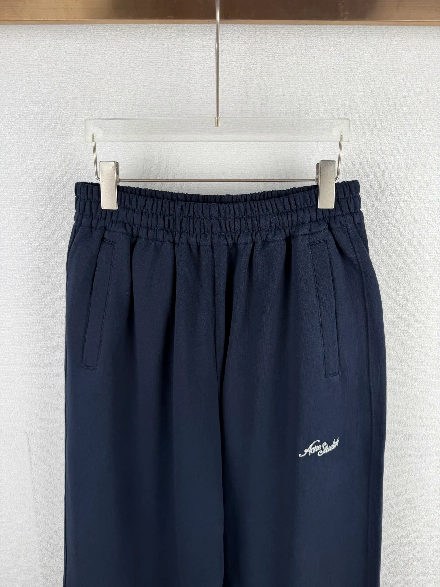 ACNE STUDIOS embroidered logo fleece sweatpants wide-leg trousers. Fabric: 32-count double yarn top + 16-count terry bottom yarn G The veil is made of 32-count fine yarn. The high-count yarn is smooth and delicate. After weaving, it forms a smooth and compact surface. It not only resists pilling and wear-resistant, but also gives the fabric a touch as delicate as skin. The base yarn is made of 16-count roving. The low-count yarn is fluffy and hairy. It naturally forms a rich terry layer during the weaving process, providing a sufficient source of fiber for subsequent sanding. This combination of fine surfaces and thick soles strikes a perfect balance between warmth and skin-friendliness. Mechanical sanding is the soul process of this fabric. By rubbing the bottom surface of the roller wrapped with emery leather at high speed, the fiber ends of the 16-count roving are evenly pulled out and cut off, forming a dense and uniform short pile layer. The smoothness and neatness of needle embroidery and the soft velvet feel of brushed fabrics form a material dialogue between hardness and softness. The embroidery area is smooth and crisp, with sharp lines, like delicate inscriptions carved on a rich suede canvas; while the surrounding brushed fabric presents a soft matte natural texture. The spacing between stitches is consistent and the strength is balanced to ensure that each stitch is placed accurately. This rigorous stitching structure makes the edges of the logo present a sharp and clear outline, and the strokes of the text are full and smooth. Even the curved turning points of the floral fonts can achieve a smooth transition through the connection of tight stitches.